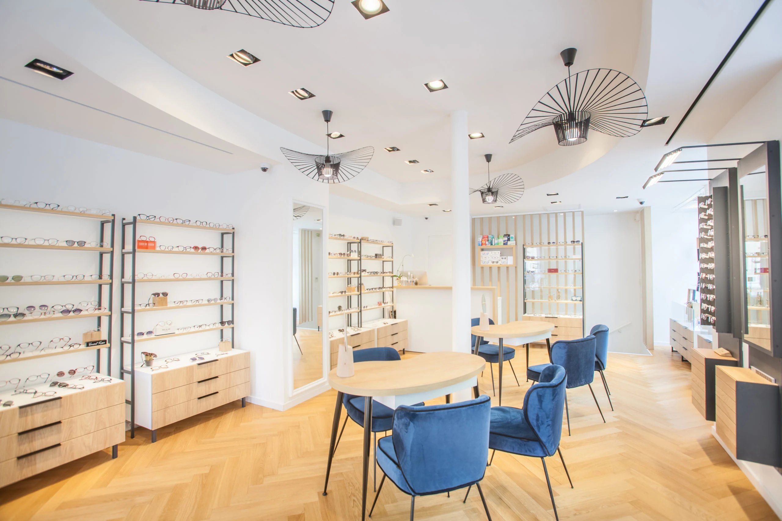 Intérieur de CONSTANT Opticiens, parquet chevron chêne, fauteuils bleus, luminaires Vertigo et linéaires de montures
